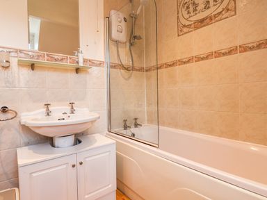 A bathroom with a white sink and cabinet, a wall-mounted mirror and shelf, a bathtub with a glass shower screen, and beige tiled walls at Lonsdale Villa Scarborough