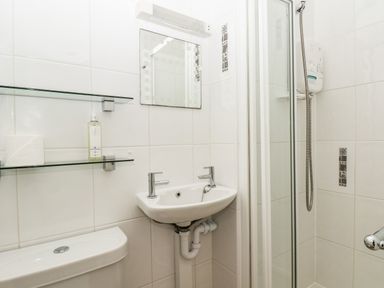 A bathroom with a toilet, small sink with two taps, a mirror, glass shelves holding a toilet paper roll and a bottle, and a shower with a sliding glass door at Lonsdale Villa in Scarborough