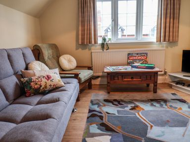 A living room with a gray sofa and patterned cushions a wooden armchair a wooden coffee table with a Monopoly game on it a patterned rug and a window with curtains at Lonsdale Villa in Scarborough