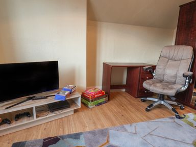A room with a TV on a stand a gaming console and controllers a stack of board games a wooden desk and an office chair at Lonsdale Villa in Scarborough