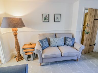 A living room with a sofa with three cushions a wooden floor lamp a set of nesting tables and a wooden door at Blossom Cottage in Broadway