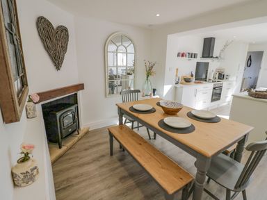 A dining area with a wooden table and chairs next to a small fireplace and a kitchen in the background at Blossom Cottage in Broadway