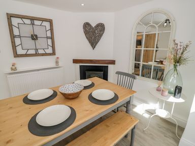 A dining area with a wooden table set with four white plates and a bowl a large wall clock a heart-shaped decoration above a fireplace and a mirror and flowers on a side table at Blossom Cottage in Broadway