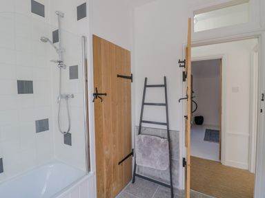 A bathroom with a bathtub and shower head wooden doors and a black towel ladder with a towel at Blossom Cottage in Broadway