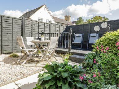 An outdoor patio area with a wooden table and chairs on gravel and a deck with two reclining chairs at Blossom Cottage in Broadway