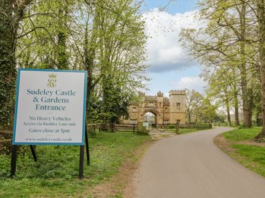 An entrance sign for Sudeley Castle and Gardens next to a road leading to a stone gatehouse surrounded by trees