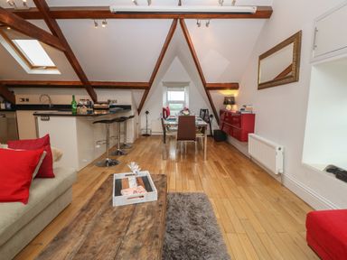 An open plan living and dining area with wooden beams a wooden floor a kitchen island with bar stools a dining table with chairs and a sofa with red cushions at Apartment 2 Gorwel Glas in Llanbedrog