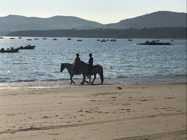 Two people riding horses on a sandy beach with boats on the water and hills in the background at Apartment 2, Gorwel Glas in Llanbedrog