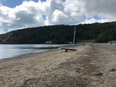 A sandy beach with a small sailboat on the shore and forested hills in the background at Apartment 2 Gorwel Glas in Llanbedrog