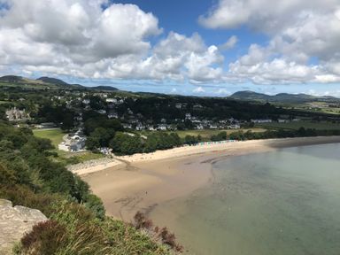 A sandy beach with colorful beach huts along the shore and a village with houses and trees in the background at Apartment 2 Gorwel Glas in Llanbedrog