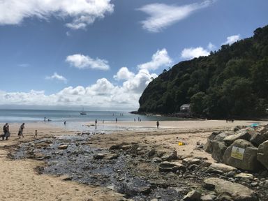 A sandy beach with people walking and a stream flowing towards the sea with a hill covered in trees at Apartment 2 Gorwel Glas in Llanbedrog