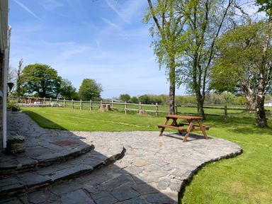 A stone patio with a wooden picnic table near trees and a grassy yard with a wooden fence at The Stable @ Canllefaes in Penparc near Cardigan