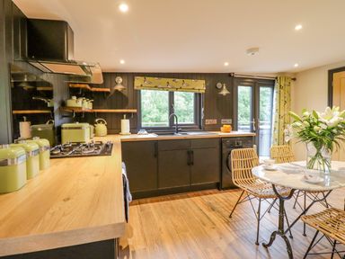 A kitchen with wooden countertop, black cabinets, wicker chairs at a round table with a vase of flowers and windows with yellow bird curtains at The Cabin at Evelyns Retreat in Glyndyfrdwy