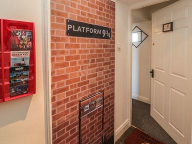 An indoor hallway with a brick wall decal and platform 9 3/4 sign next to a white door and a red brochure holder with maps at The Wizards Loft in Alnwick