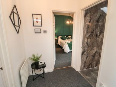 A hallway with framed pictures on the wall a small black table with a plant and a thermostat visible through the door is a bedroom with a bed and green accent wall at The Wizard's Loft in Alnwick
