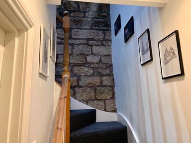 A staircase with blue carpet and wooden handrail next to a stone wall and framed pictures on walls at The Wizard's Loft in Alnwick
