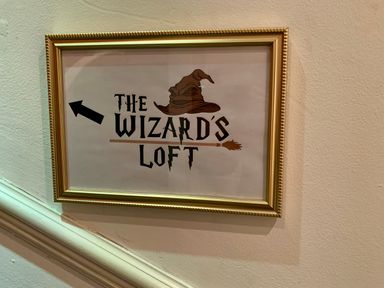 A framed sign on a wall with text The Wizard's Loft and a wizard hat above a broomstick pointing left at The Wizard's Loft in Alnwick