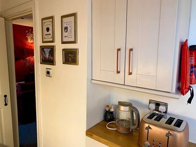 A kitchen corner with a kettle and a toaster on a wooden countertop under white cabinets with framed pictures on the wall and a fire extinguisher mounted nearby at The Wizard's Loft in Alnwick