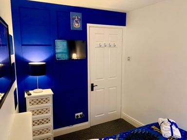 A bedroom corner with a white door a mounted TV a small white chest of drawers with a lamp and a blue accent wall at The Wizard's Loft in Alnwick