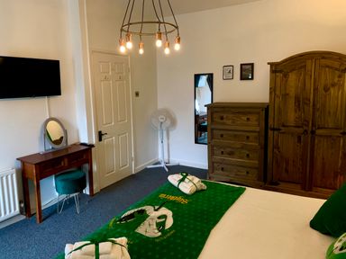 A bedroom with a bed covered with a green Hogwarts blanket a wooden dresser wooden wardrobe a vanity table with a green stool a wall-mounted TV and a standing fan at The Wizards Loft in Alnwick