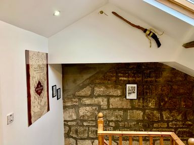 A staircase area with stone wall and wooden railing with a broomstick mounted on the wall and framed pictures at The Wizards Loft in Alnwick