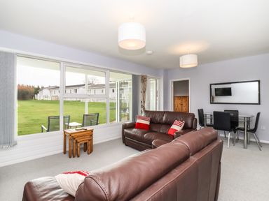 A living room with brown leather sofas red and white cushions a wooden nested table large windows and a dining table with black chairs at Marsh Bungalow in Lowestoft