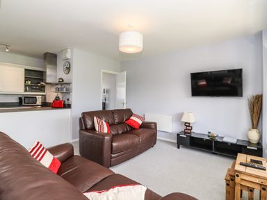 A living room with brown leather sofas pillows a wall-mounted TV a black cabinet and an open kitchen at Marsh Bungalow in Lowestoft