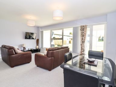 A living room with brown leather sofas a wall-mounted television a glass dining table with black chairs and large windows with curtains at Marsh Bungalow in Lowestoft