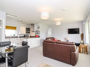 An open plan living room and kitchen with brown leather sofas a glass dining table and white kitchen cabinets at Marsh Bungalow in Lowestoft