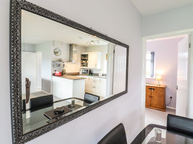 A dining area with a glass table and black chairs facing a large decorative wall mirror reflecting a kitchen with white cabinets and appliances at Marsh Bungalow in Lowestoft