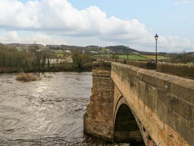 A stone bridge over a flowing river with lampposts along the bridge and fields in the background at Bramwell House in Corbridge