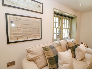 A sitting area with a beige sofa and patterned cushions under a window with two vases and framed documents on the wall at Bramwell House in Corbridge