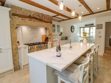A kitchen with an island countertop with chairs and a bottle with glasses near a stove set in a stone fireplace at Bramwell House in Corbridge