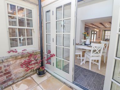 A patio with a potted plant outside glass doors leading to a dining area with white chairs and a table with flowers at Bramwell House in Corbridge