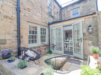 A stone patio with two black chairs a bottle of champagne and glasses and potted plants outside a house with open glass doors at Bramwell House in Corbridge