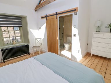 A bedroom with a bed a white chair a window a radiator a chest of drawers and an en suite bathroom with a wooden sliding door at Bramwell House in Corbridge