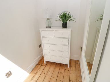A white chest of drawers with a lamp and a potted plant on top in a corner with wooden floor and a full-length mirror