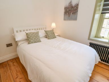 A bedroom with a white bed and two patterned pillows a bedside table with a lamp a window and wooden floor at Bramwell House in Corbridge