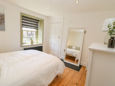 A bedroom with a bed white bedding a window with a striped blind a mirror leaning against the wall a door and a white dresser with decorative items at Bramwell House Corbridge