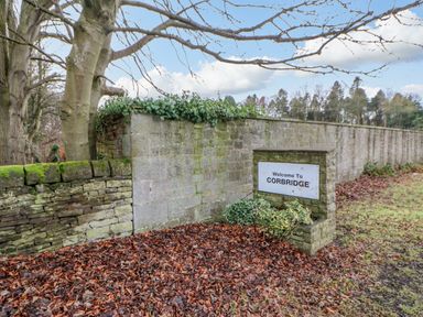A stone wall with a welcome to Corbridge sign and fallen leaves on the ground at Bramwell House in Corbridge