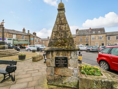 A stone monument with a plaque in a town square with benches parked cars and stone buildings with shops in the background at Bramwell House in Corbridge