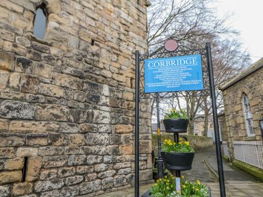 A stone wall and a blue historical sign about Corbridge next to flower pots on a paved street at Bramwell House in Corbridge