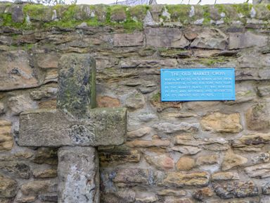 An old stone market cross mounted on a stone wall with a blue information plaque at Bramwell House in Corbridge