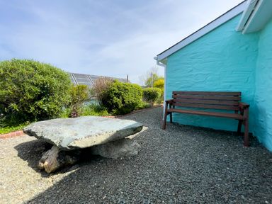 A wooden bench and a large flat stone table on a gravel ground beside a turquoise painted wall with bushes in the background at Ty Coed @ Canllefaes in Penparc near Cardigan