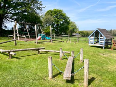 An outdoor playground with wooden balance beams swings a small playhouse and a climbing slide at Ty Coed @ Canllefaes in Penparc near Cardigan