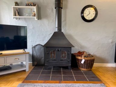 A living room with a black wood stove on a tiled hearth a wall clock a TV on a stand and a basket with firewood at The Granary @ Canllefaes in Penparc near Cardigan
