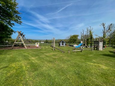 A playground with swings a slide a climbing structure and a wooden playhouse on grass at The Granary @ Canllefaes in Penparc near Cardigan