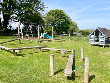 An outdoor playground with wooden play equipment and a small playhouse on grass at The Granary @ Canllefaes Penparc near Cardigan