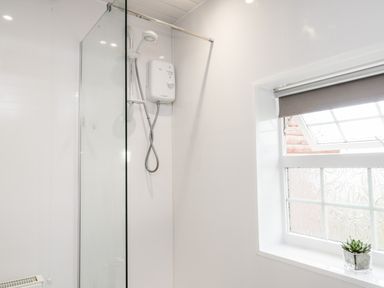 A shower area with a glass partition an electric shower unit and a window with a grey roller blind and a small plant on the sill at Hawthorn Dale Cottage Whitby