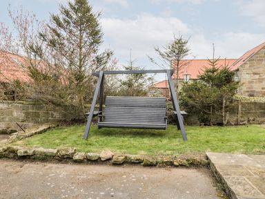 A garden with a black wooden swing on a grassy area with stone walls and small trees at Hawthorn Dale Cottage in Whitby
